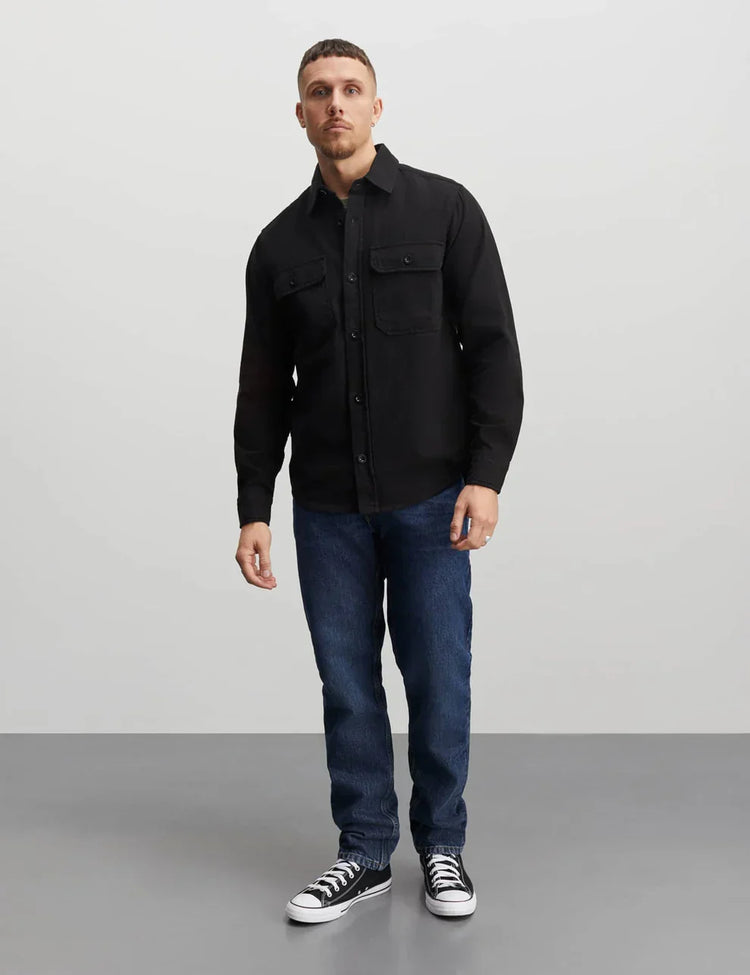 Man i svart skjortjacka, blå jeans och svarta sneakers mot ljus bakgrund.