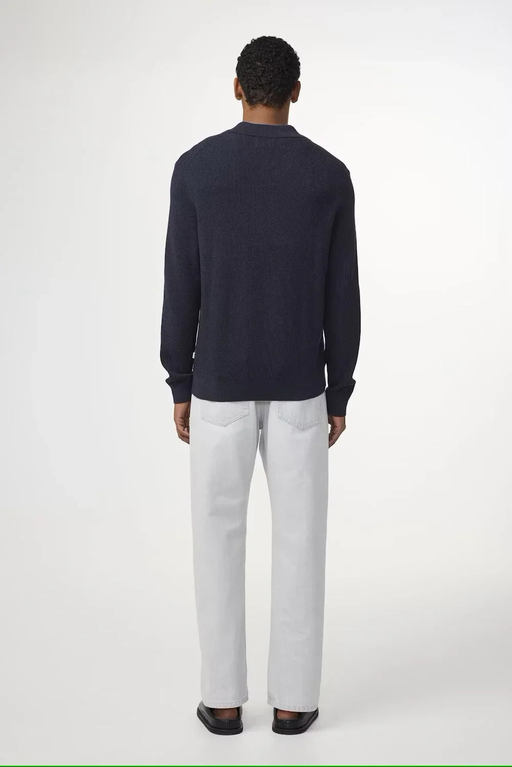 Man i mörkblå stickad tröja och ljusa raka jeans, sedd bakifrån mot vit bakgrund