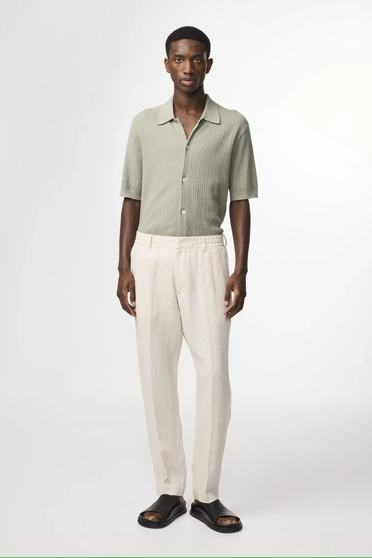 Man i ljusgrön stickad skjorta, beige linnebyxor och svarta sandaler mot vit bakgrund