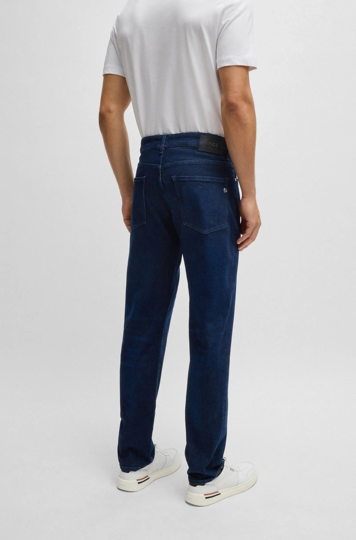 Man i vita sneakers och vita t-shirt bär blå herrjeans, sedd bakifrån, mot ljus bakgrund.