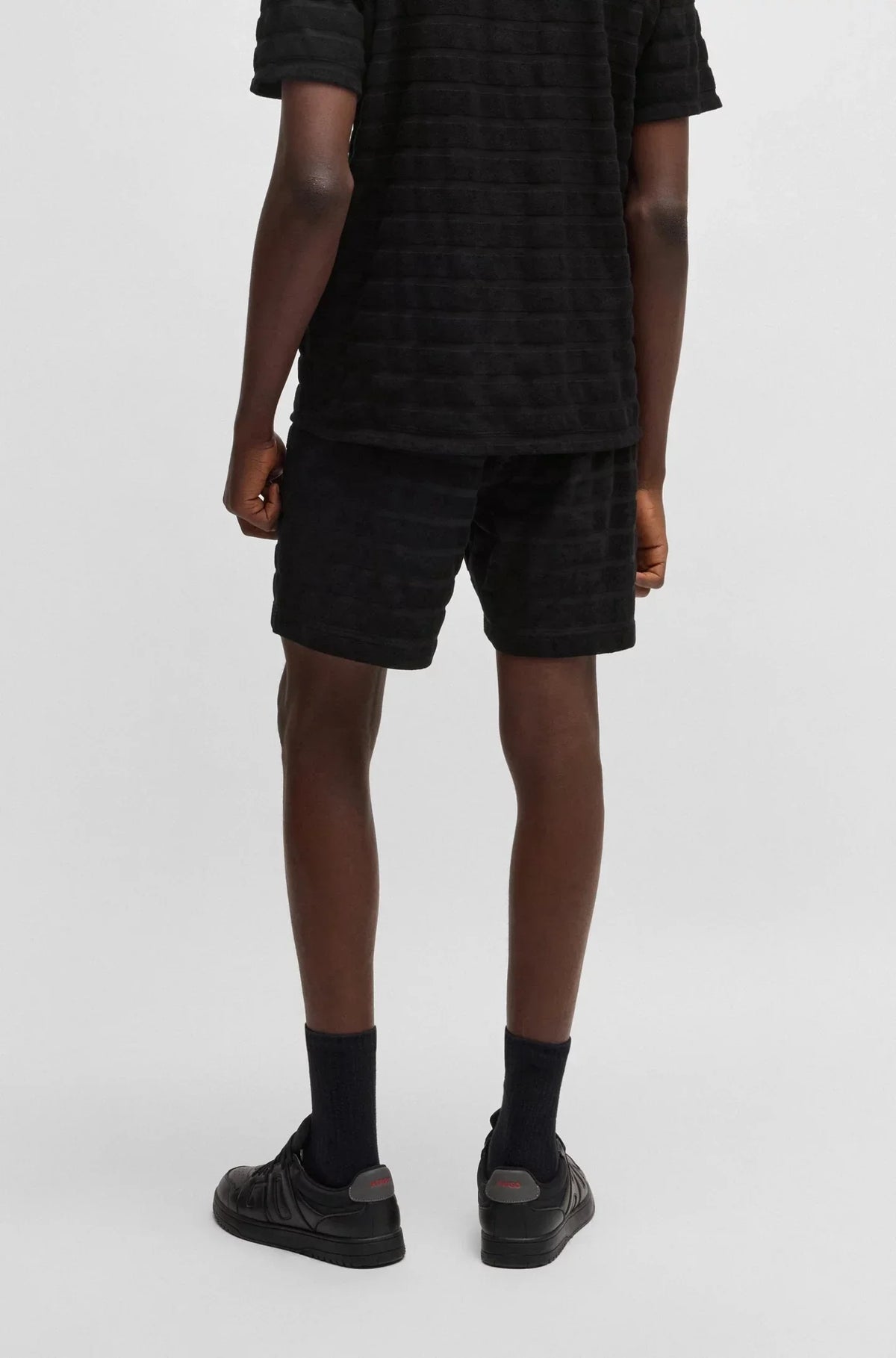 Herr i svart kortärmad tröja och matchande svarta shorts, bakifrån, mot vit bakgrund.