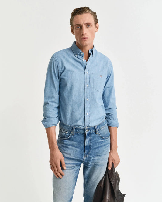 Man i ljusblå jeansskjorta och blå jeans, håller en mörk jacka, mot vit bakgrund.