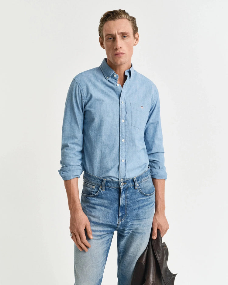 Man i ljusblå jeansskjorta och blå jeans, håller en mörk jacka, mot vit bakgrund.