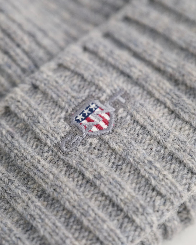 Gråstickad herrmössa med broderad sköld i fokus, nära detaljbild