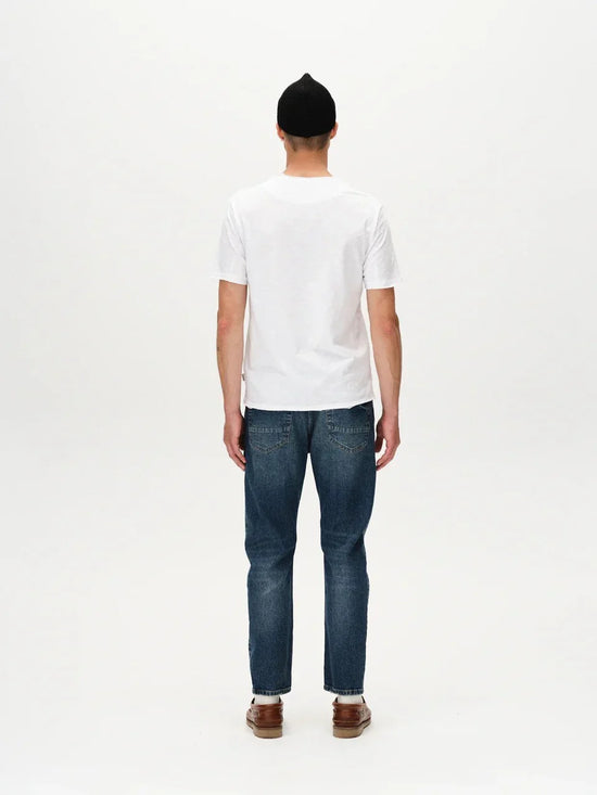 Man i vit t-shirt, blå jeans och bruna skor sedd bakifrån mot vit bakgrund