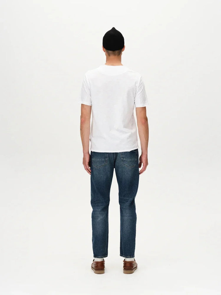Man i vit t-shirt, blå jeans och bruna skor sedd bakifrån mot vit bakgrund