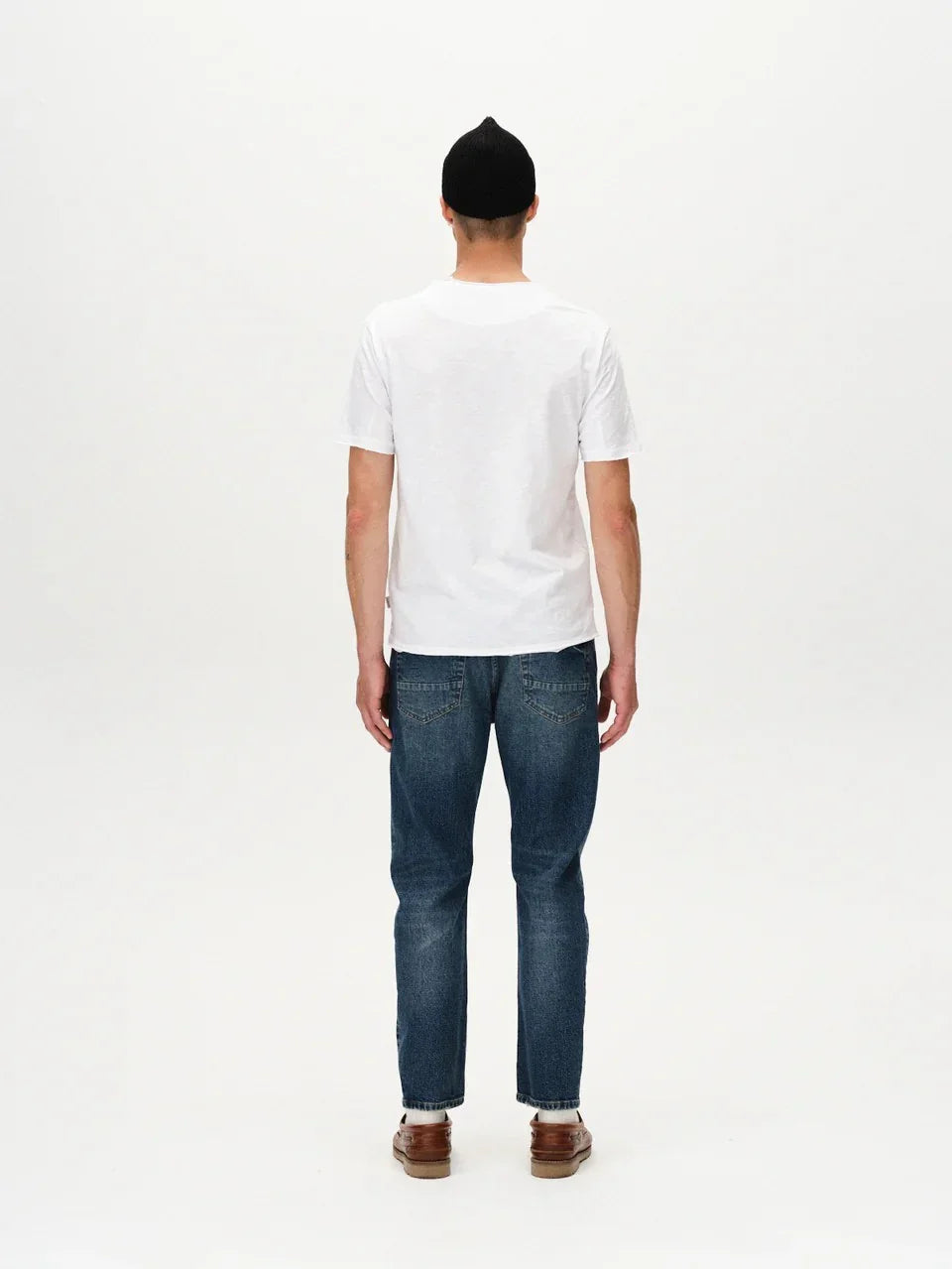 Man i vit t-shirt, blå jeans och bruna skor sedd bakifrån mot vit bakgrund