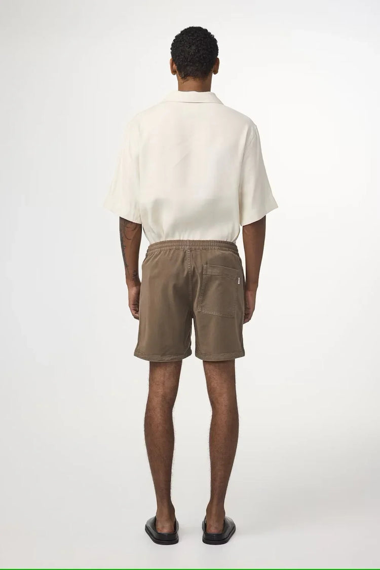Herrmodell bakifrån med beige skjorta, bruna kortbyxor och sandaler mot vit bakgrund