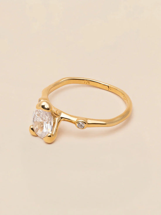 Elegant guldring med oval klar sten och liten rund diamant på ljus bakgrund