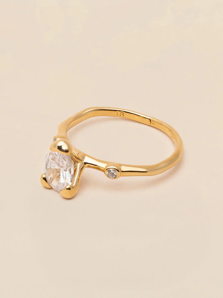 Elegant guldring med oval klar sten och liten rund diamant på ljus bakgrund