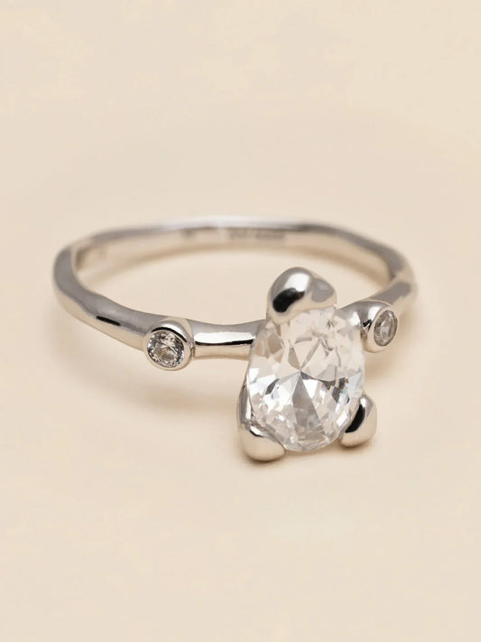 Elegant silverring med stor dropformad klar sten och två små runda stenar mot ljus bakgrund.