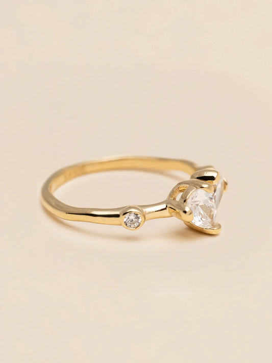 Elegant guldring med hjärtformad klar sten och liten diamant på beige bakgrund