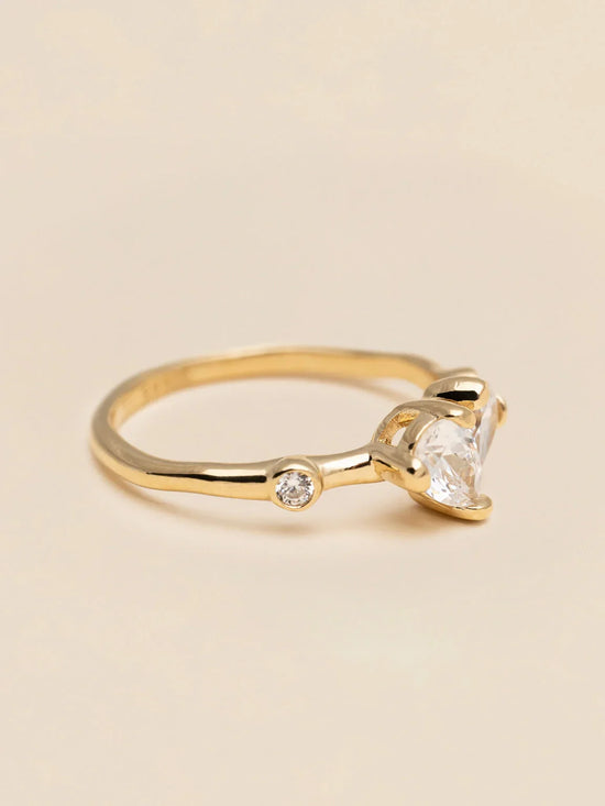 Elegant guldring med hjärtformad klar sten och liten diamant på beige bakgrund