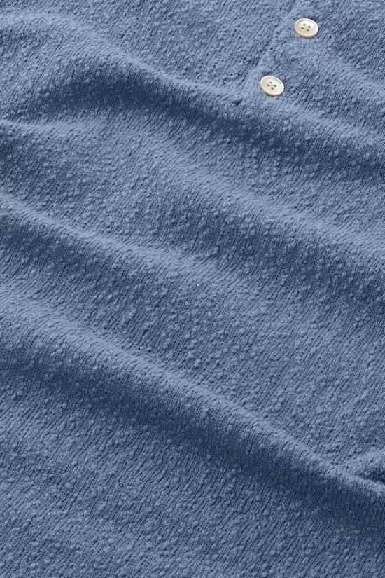 Närbild på blå pikétröja i texturerat tyg med två ljusa knappar, klassisk herrstil