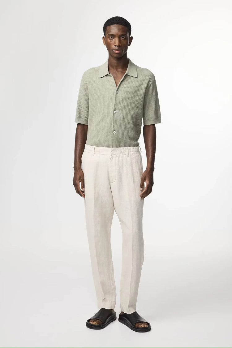 Man i ljusgrön stickad skjorta, beige linnebyxor och svarta sandaler mot vit bakgrund