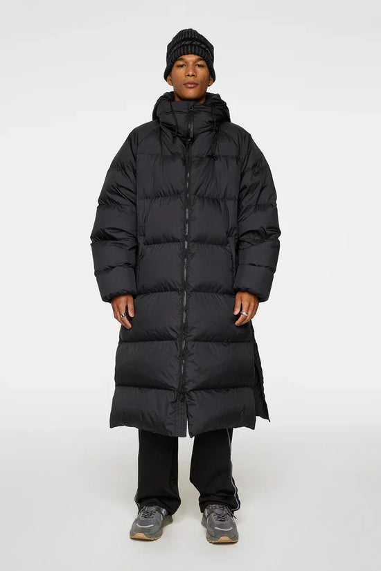 Man i lång svart dunjacka, mössa och sneakers, stående mot vit bakgrund.