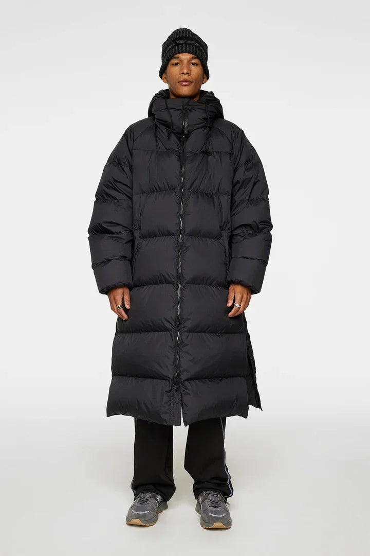 Man i lång svart dunjacka, mössa och sneakers, stående mot vit bakgrund.