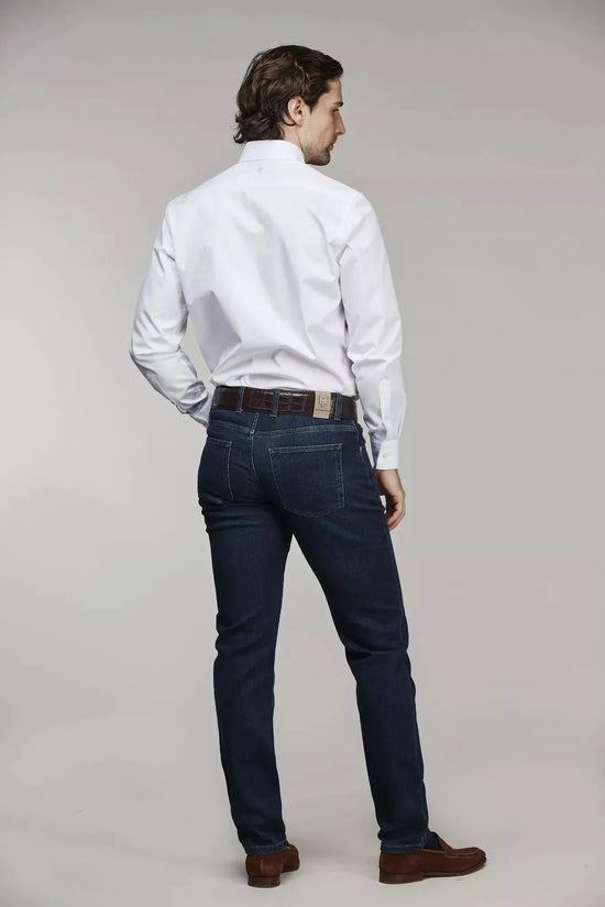 Man i vit skjorta och mörkblå denimbyxor med brunt bälte, fotad bakifrån mot grå bakgrund.