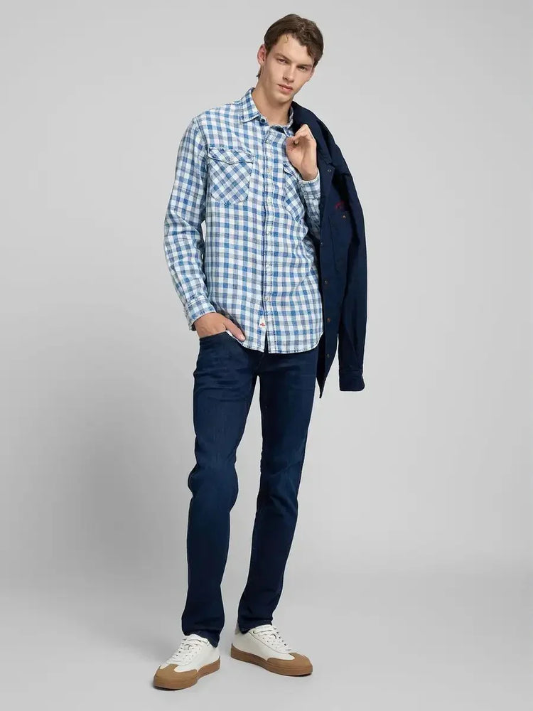 Man i blåvitrutig skjorta och mörka jeans, vita sneakers, håller en mörk jacka över axeln.