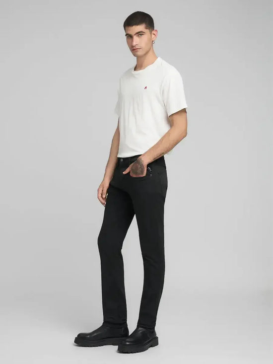 Man i vit t-shirt och svarta jeans, stående mot grå bakgrund, herrmode