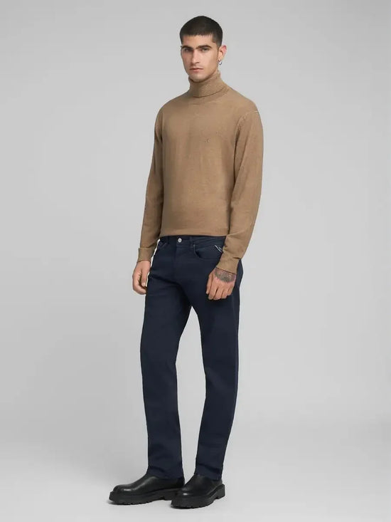 Man i beige polotröja och mörkblå byxor, stående mot ljus bakgrund, herrmode