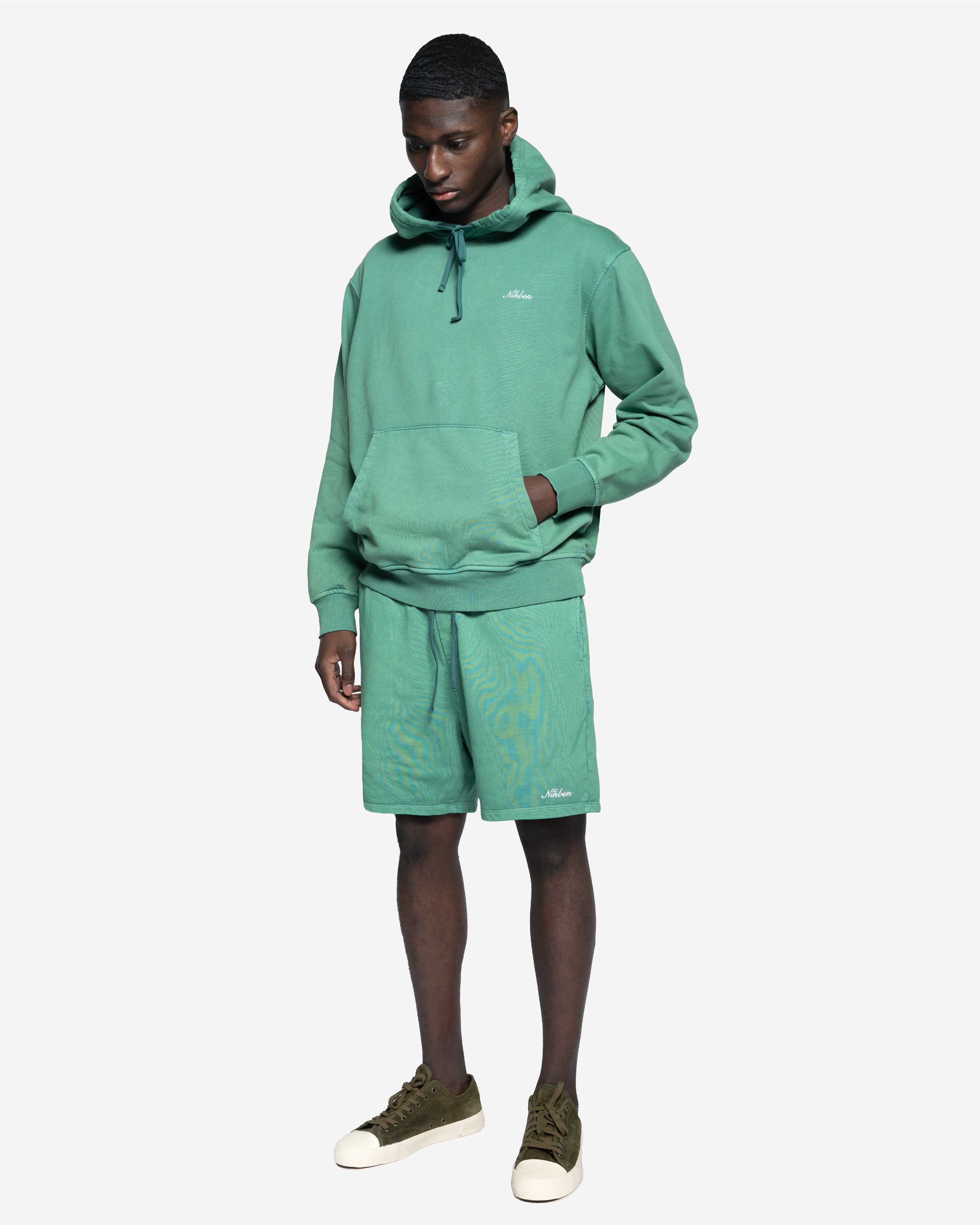 Man i grön hoodie och matchande shorts, avslappnad herrklädstil, vit bakgrund