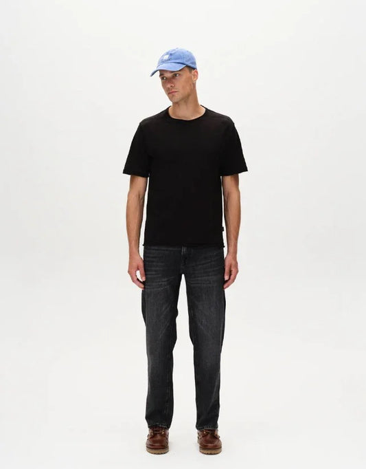 Man i svart t-shirt, blå keps och mörka jeans mot vit bakgrund