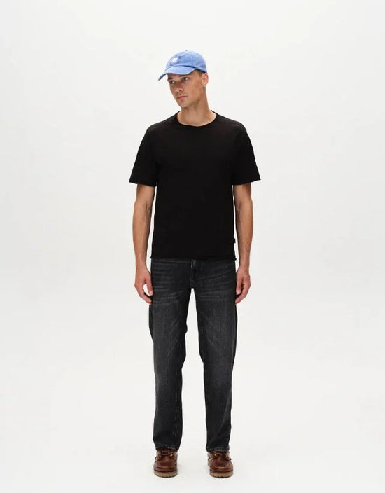 Man i svart t-shirt, blå keps och mörka jeans mot vit bakgrund