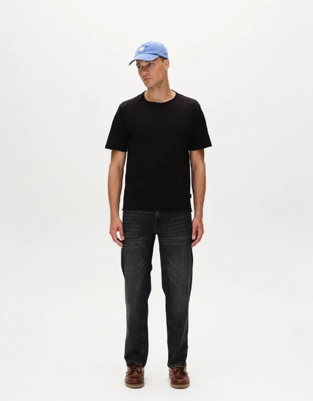 Man i svart t-shirt, blå keps och mörka jeans mot vit bakgrund