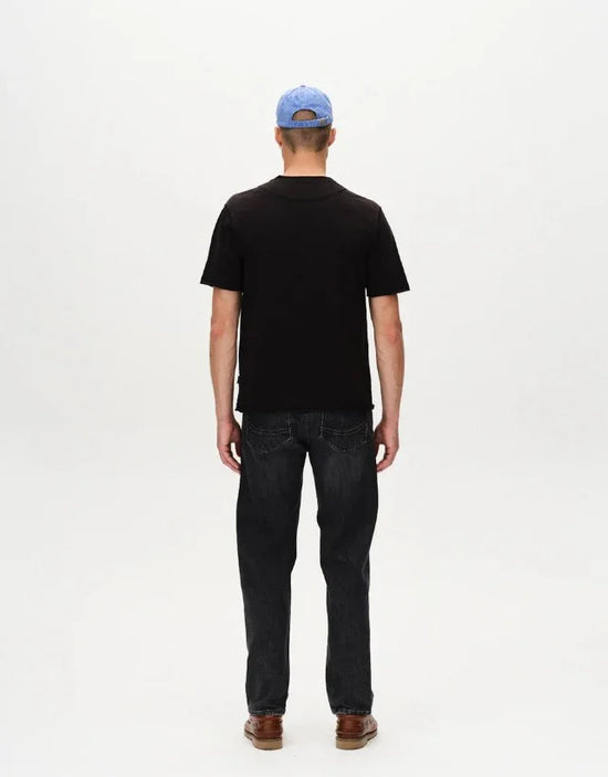 Man i svart t-shirt, mörka jeans och blå keps fotad bakifrån mot vit bakgrund