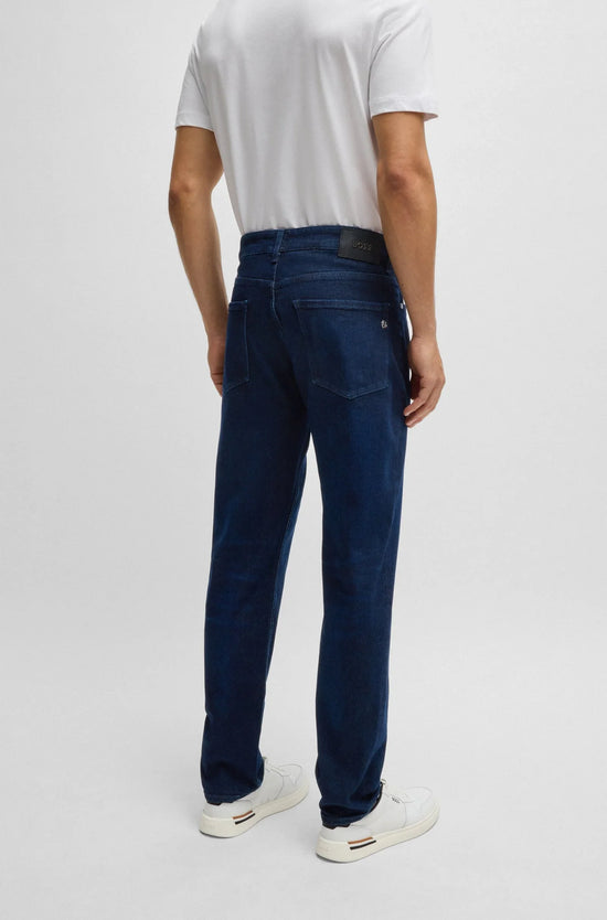 Man i vita sneakers och vita t-shirt bär blå herrjeans, sedd bakifrån, mot ljus bakgrund.