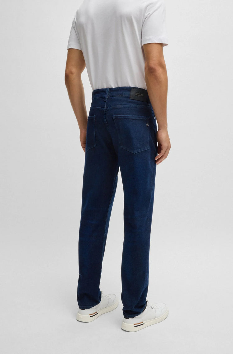Man i vita sneakers och vita t-shirt bär blå herrjeans, sedd bakifrån, mot ljus bakgrund.