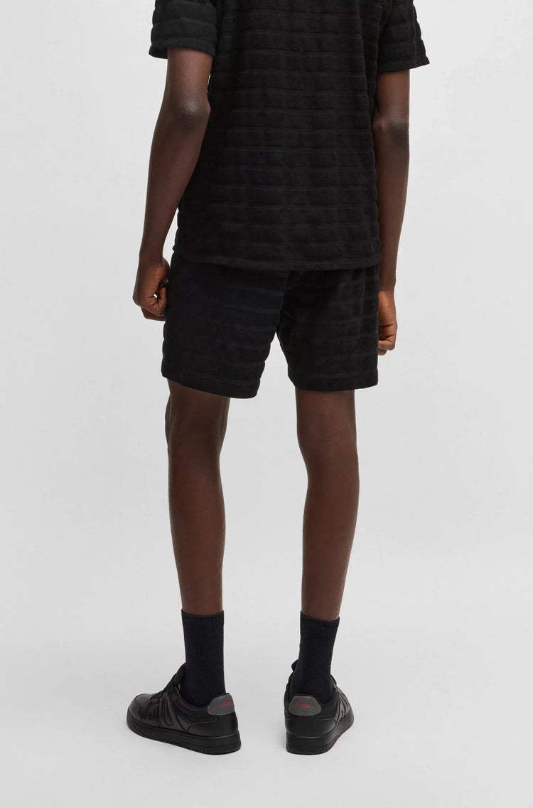 Herr i svart kortärmad tröja och matchande svarta shorts, bakifrån, mot vit bakgrund.