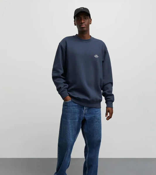 Man i mörkblå sweatshirt, jeans och svart keps mot ljus bakgrund, herrmode