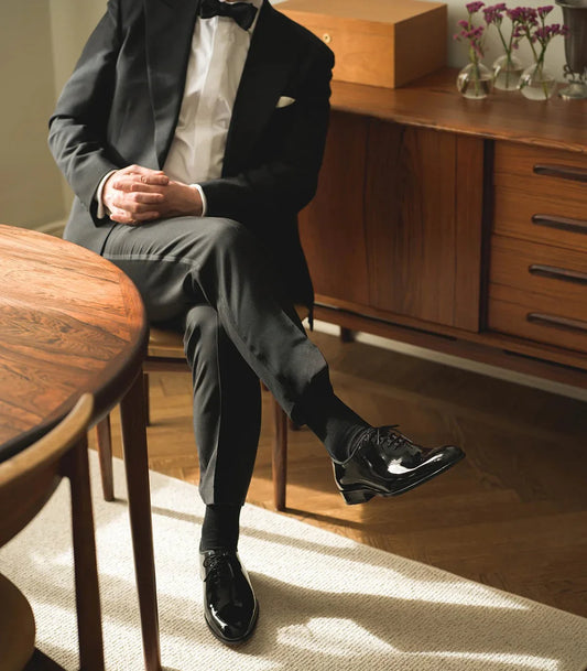 Man i svart smoking och lackskor sitter vid matbord, stilfull herrklädsel i elegant miljö.