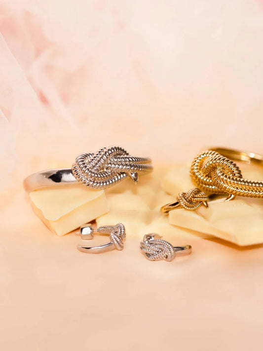 Smycken i silver och guld med knutdesign, armband och ring på beige bakgrund.