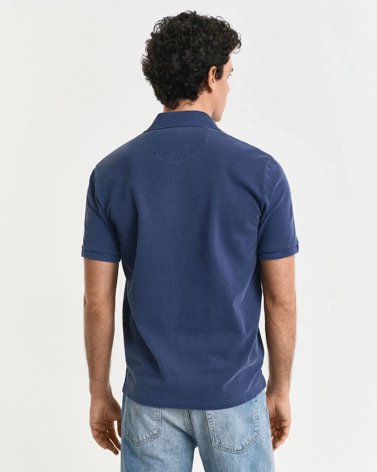 Man i blå pikétröja och ljusa jeans, fotograferad bakifrån mot vit bakgrund.