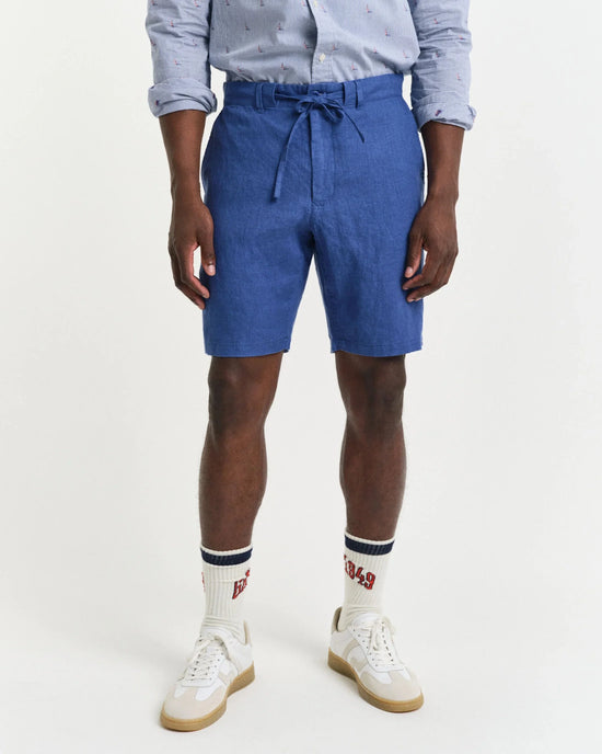 Herrkläder: man i blå linneshorts, skjorta, vita sneakers och höga strumpor mot vit bakgrund