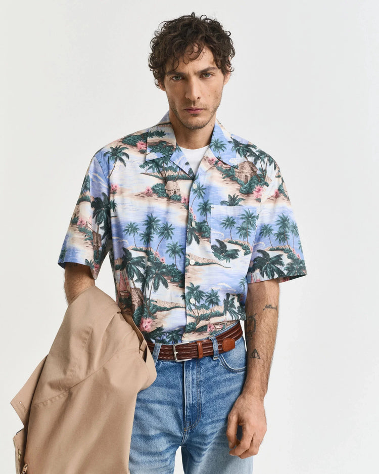 Man i kortärmad hawaiiskjorta med palmmotiv, ljusblå jeans och bälte, håller beige jacka.
