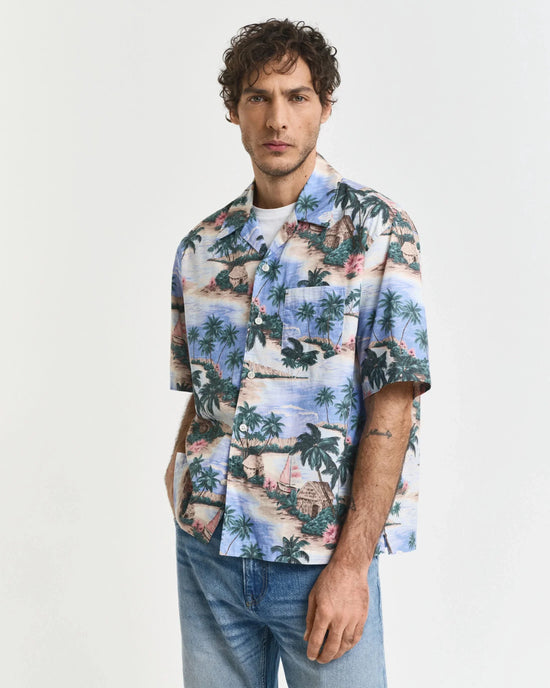 Man i mönstrad kortärmad skjorta med tropiskt motiv och blå jeans mot vit bakgrund.