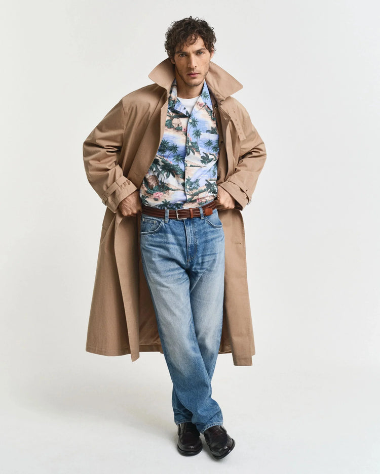 Man i beige trenchcoat, mönstrad skjorta och blå jeans mot vit bakgrund.