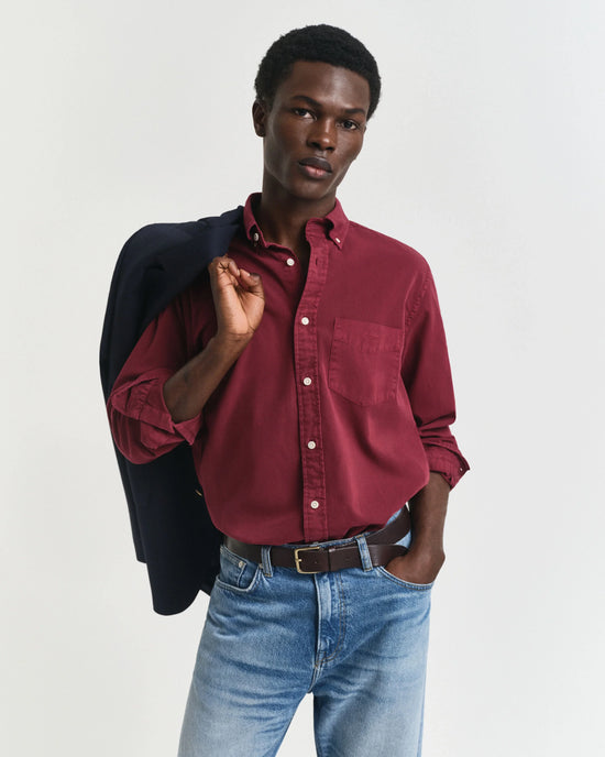 Man i vinröd skjorta och blå jeans, håller mörk kavaj över axeln, klassiskt herrmode.