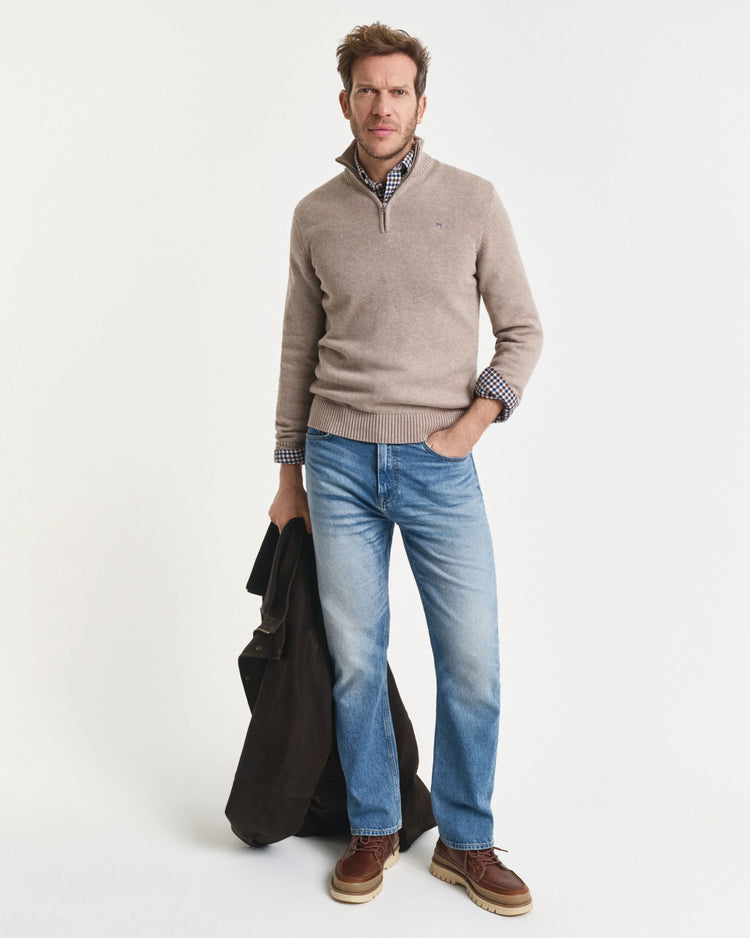 Man i beige stickad tröja, rutiga skjorta och blå jeans, håller brun jacka, herrmode