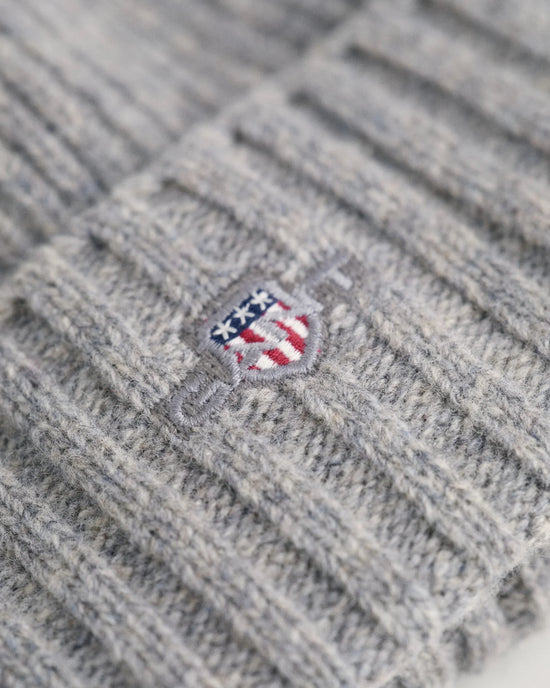 Gråstickad herrmössa med broderad sköld i fokus, nära detaljbild